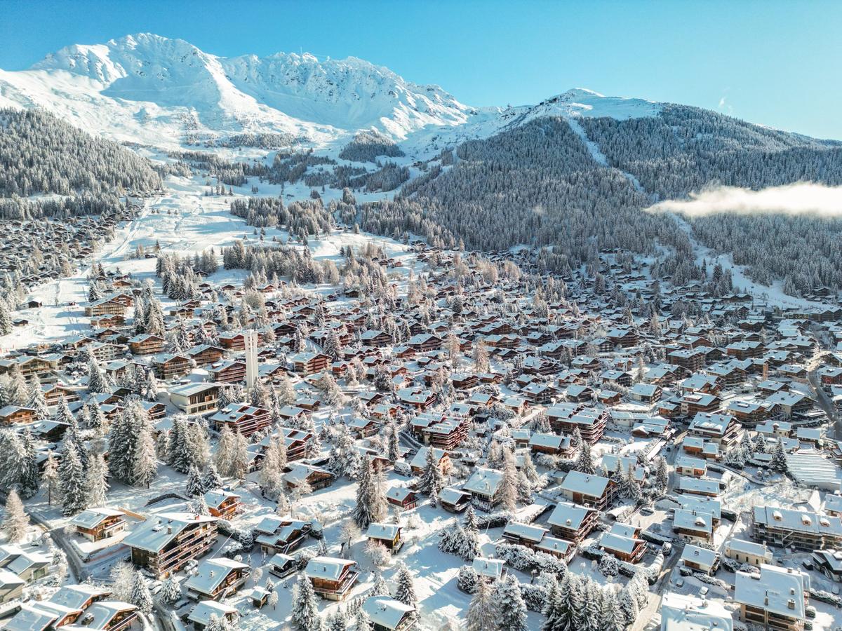Aerial view of Verbier chalets and pine trees under fresh snow, with sunlit peaks and a blue sky behind